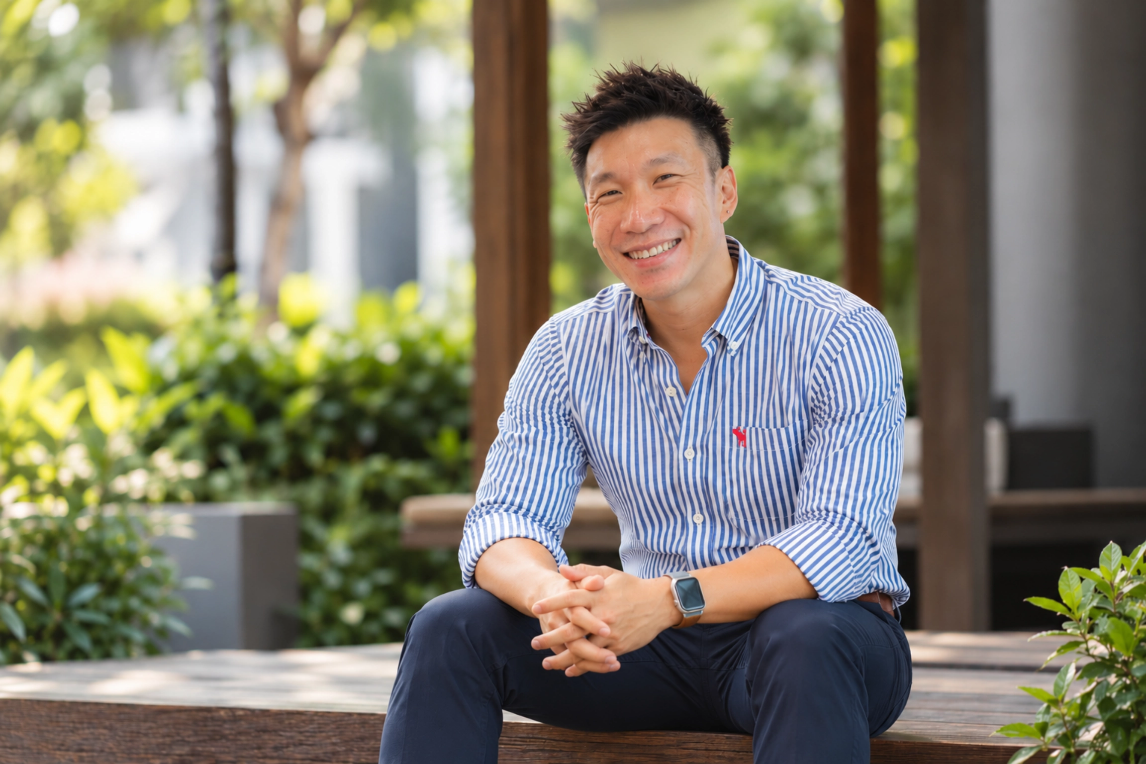 Clarry Au seated in a garden, wearing a blue-and-white striped shirt and navy trousers